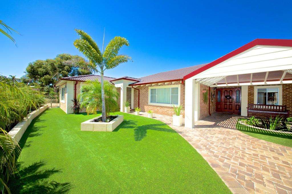 Single-story brick house with a red roof, featuring a charming granny flat. It's surrounded by artificial grass and landscaping, with a covered porch and paved pathway leading to the entrance under a clear blue Connolly sky.