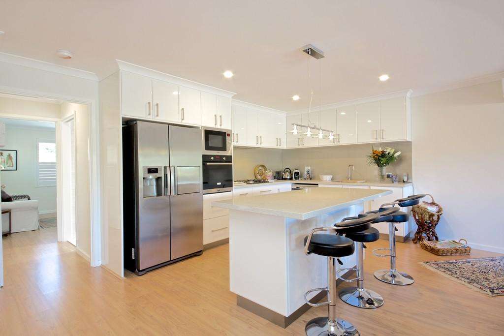 Modern kitchen with a stainless steel fridge, white cabinetry, an island with bar stools, and a hanging light fixture. The light wood flooring extends into the nearby open doorway leading to the Connolly living area in this charming granny flat.