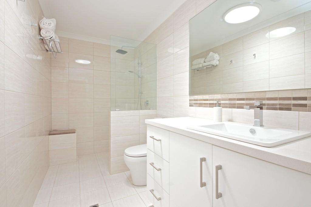Modern bathroom in a Connolly granny flat, featuring beige tiles and a glass-enclosed shower. A white vanity with a sink and toilet complements the space. Towels are neatly stacked on a wall shelf, creating an inviting ambiance.