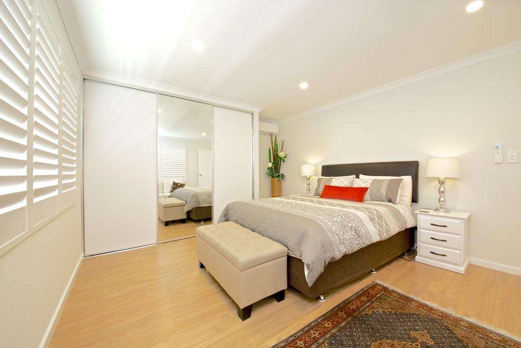 Step into this inviting bedroom, perfect for a Granny Flat, featuring a double bed with gray and white bedding adorned by a red accent pillow. Flanked by bedside tables with lamps and a large mirror door, it also offers the comfort of a beige bench at the foot of the bed.
