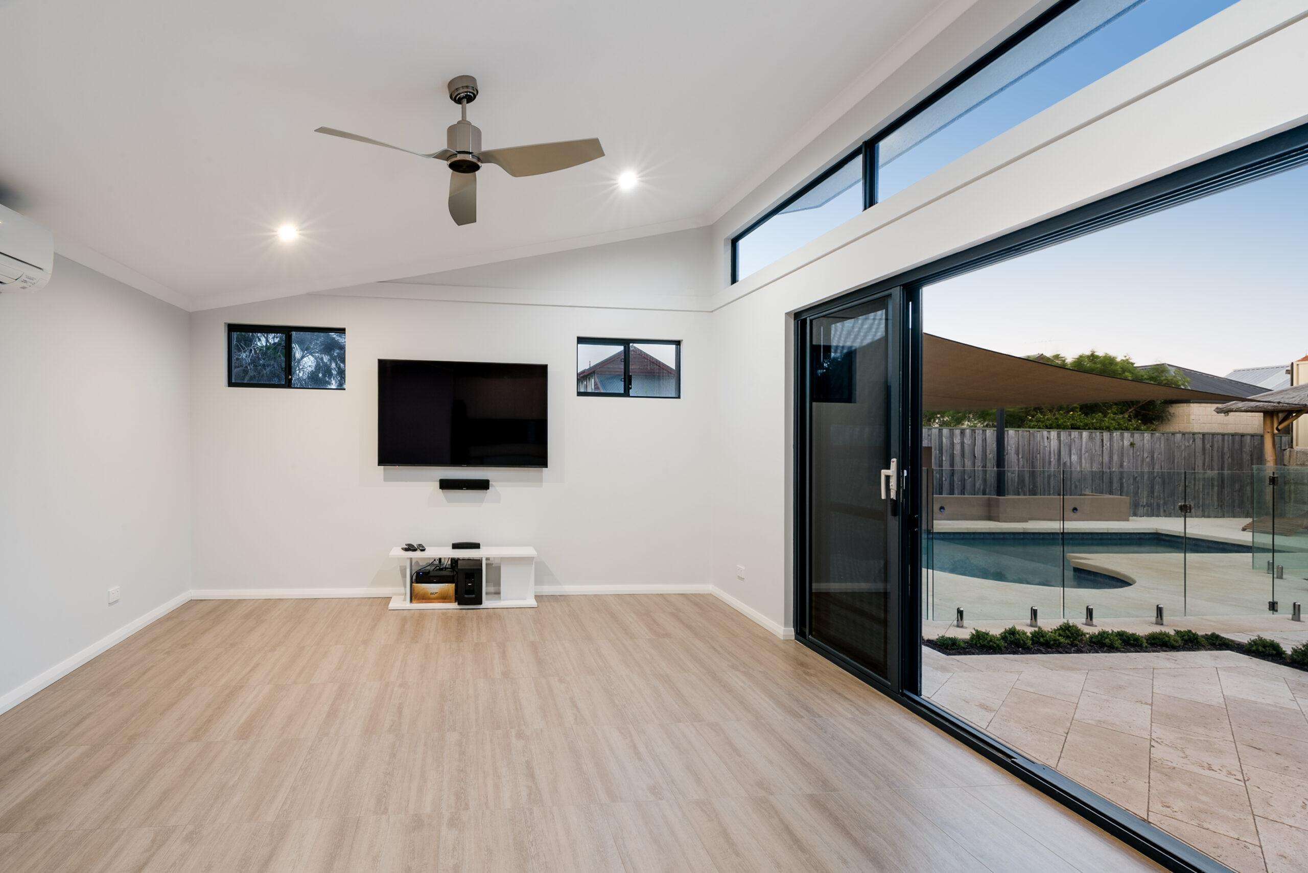 A cozy granny flat in Quinns Rocks features a minimalist living room with wooden flooring, a wall-mounted TV, and a ceiling fan. Glass doors seamlessly open to reveal a pool outside, offering both comfort and style in one elegant space.