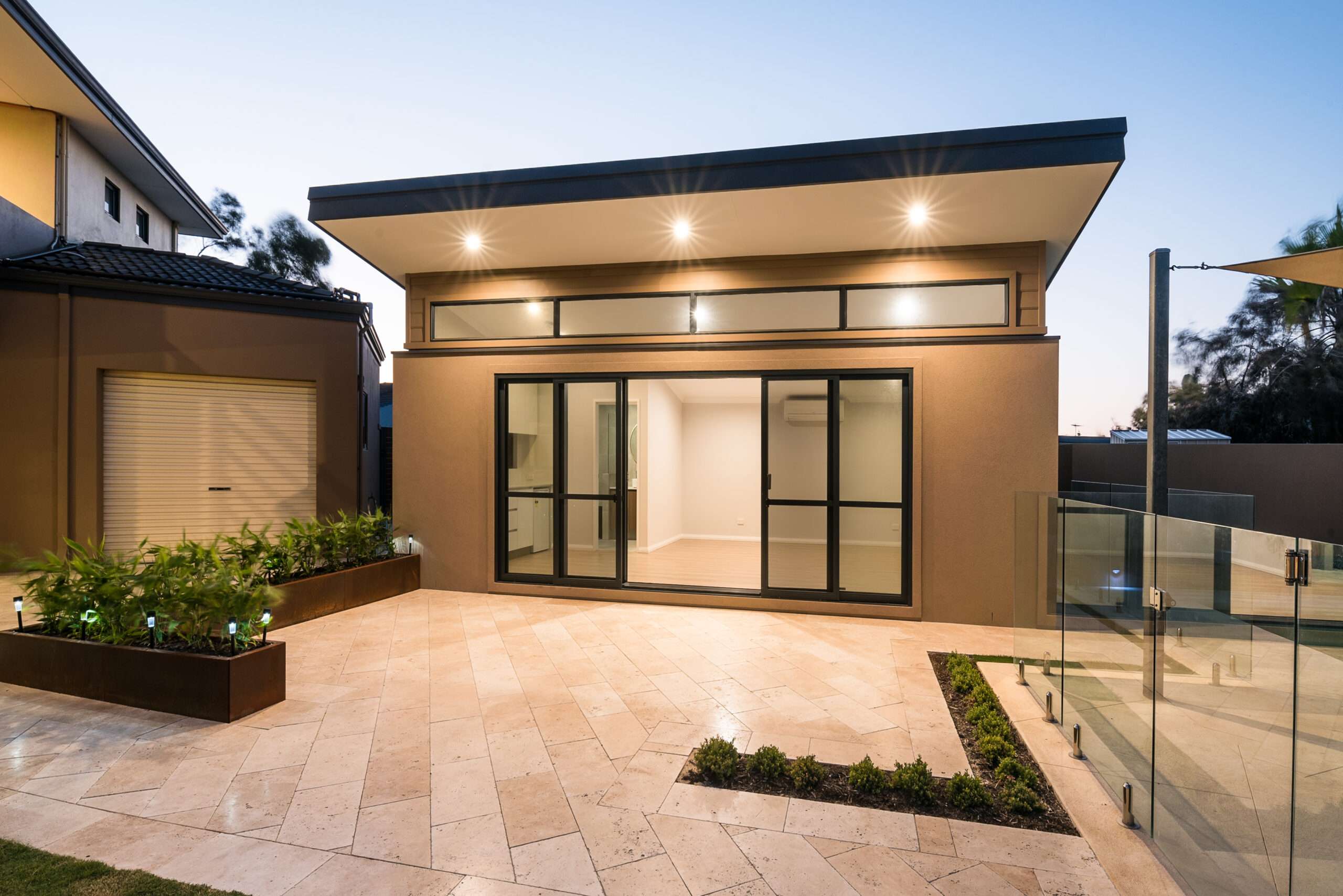 Modern single-story house with large windows and a flat roof situated in Quinns Rocks. The yard features tiled flooring, small garden beds, and a glass fence. As dusk settles, lights illuminate the exterior, offering a serene view that complements the optional granny flat perfectly.