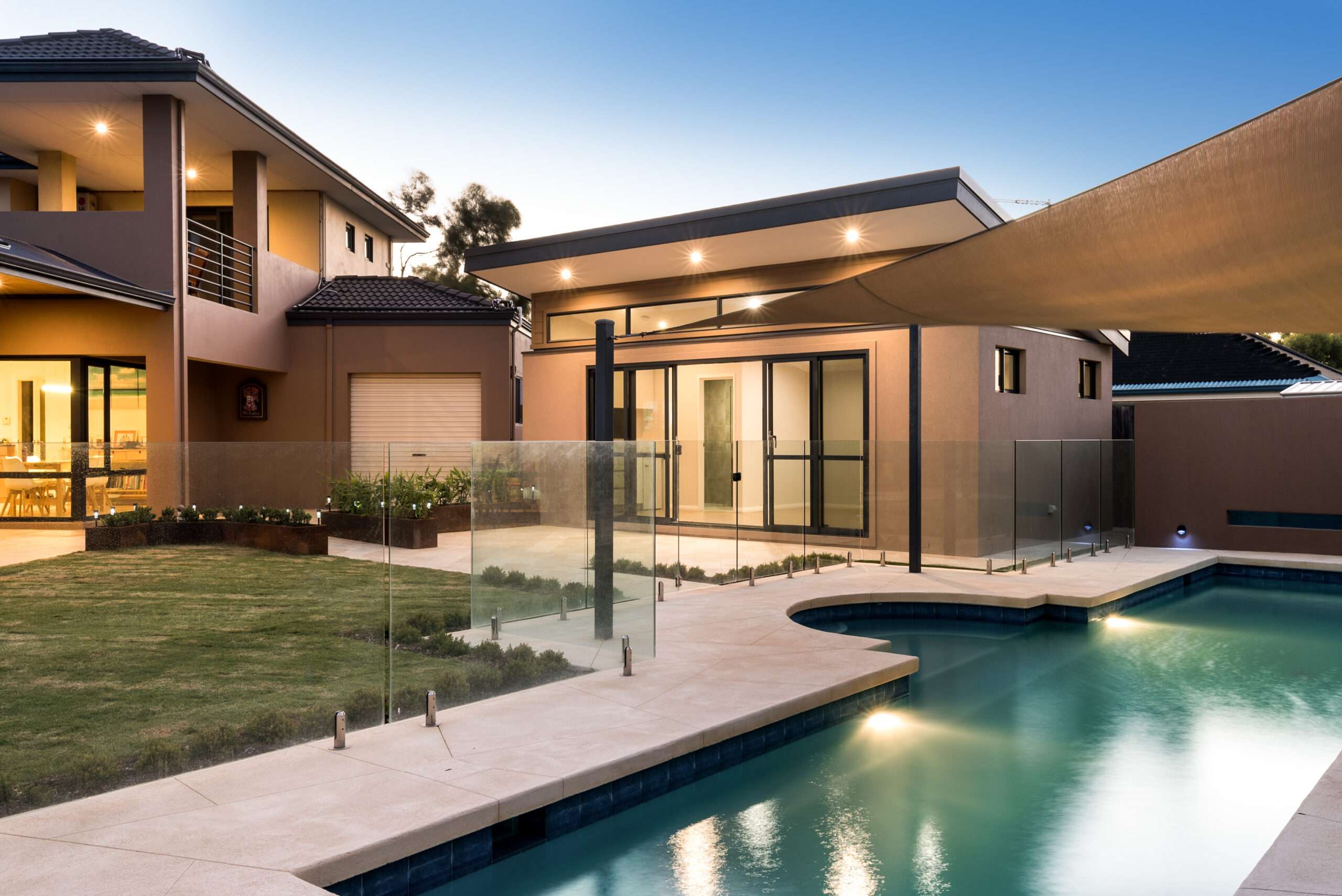 Modern house in Quinns Rocks with large windows, outdoor pool, and sleek glass fencing. Beige walls frame a well-lit patio area, while an adjacent lawn stretches under the clear blue sky. Additionally, there's a stylish Granny Flat offering extra space and comfort.