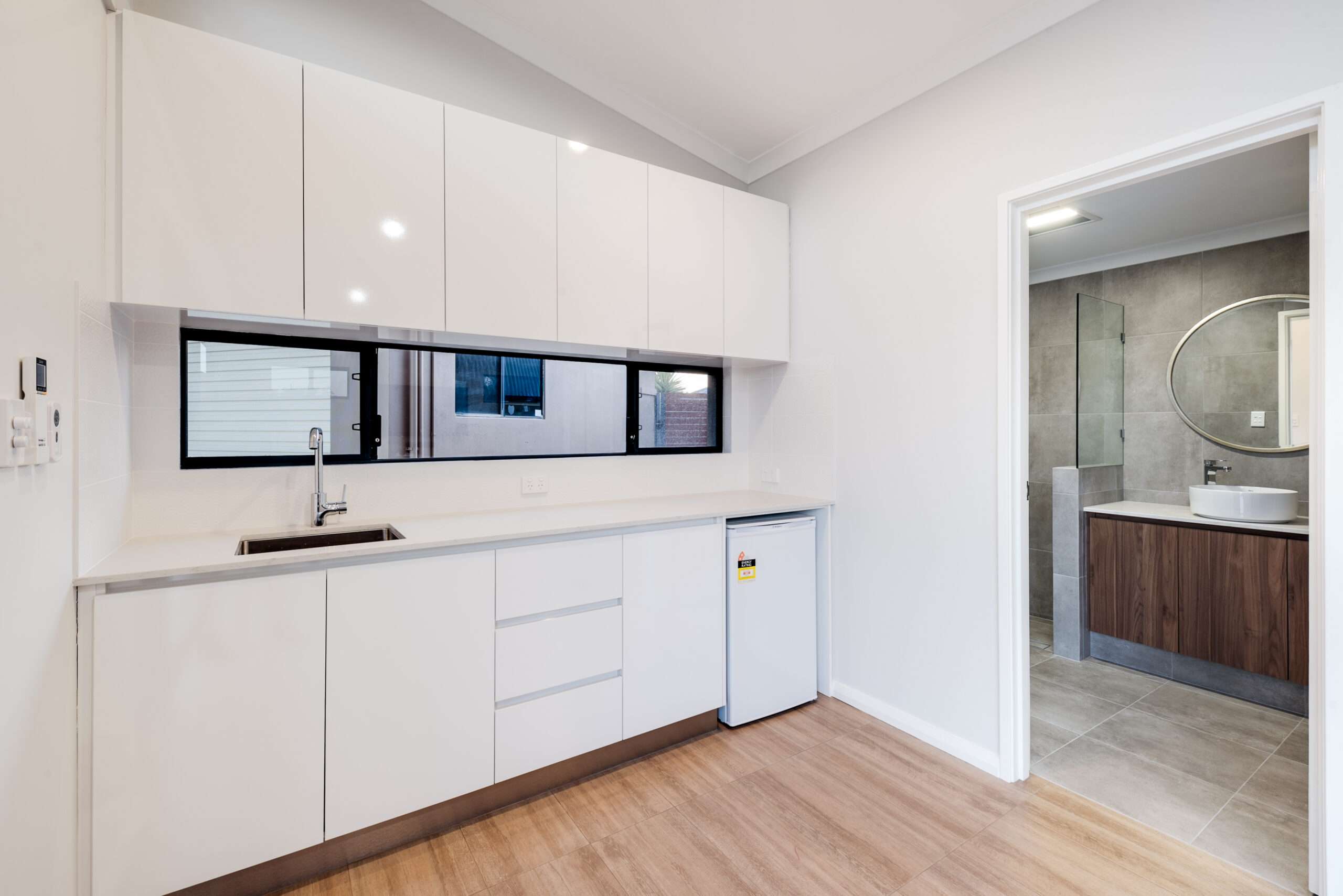 Modern kitchen with white cabinets, sink, and a mini-fridge. An open door leads to a bathroom featuring a round mirror, wooden vanity, and gray tiles. This stylish space is ideal for a Quinns Rocks granny flat, combining functionality with contemporary charm.