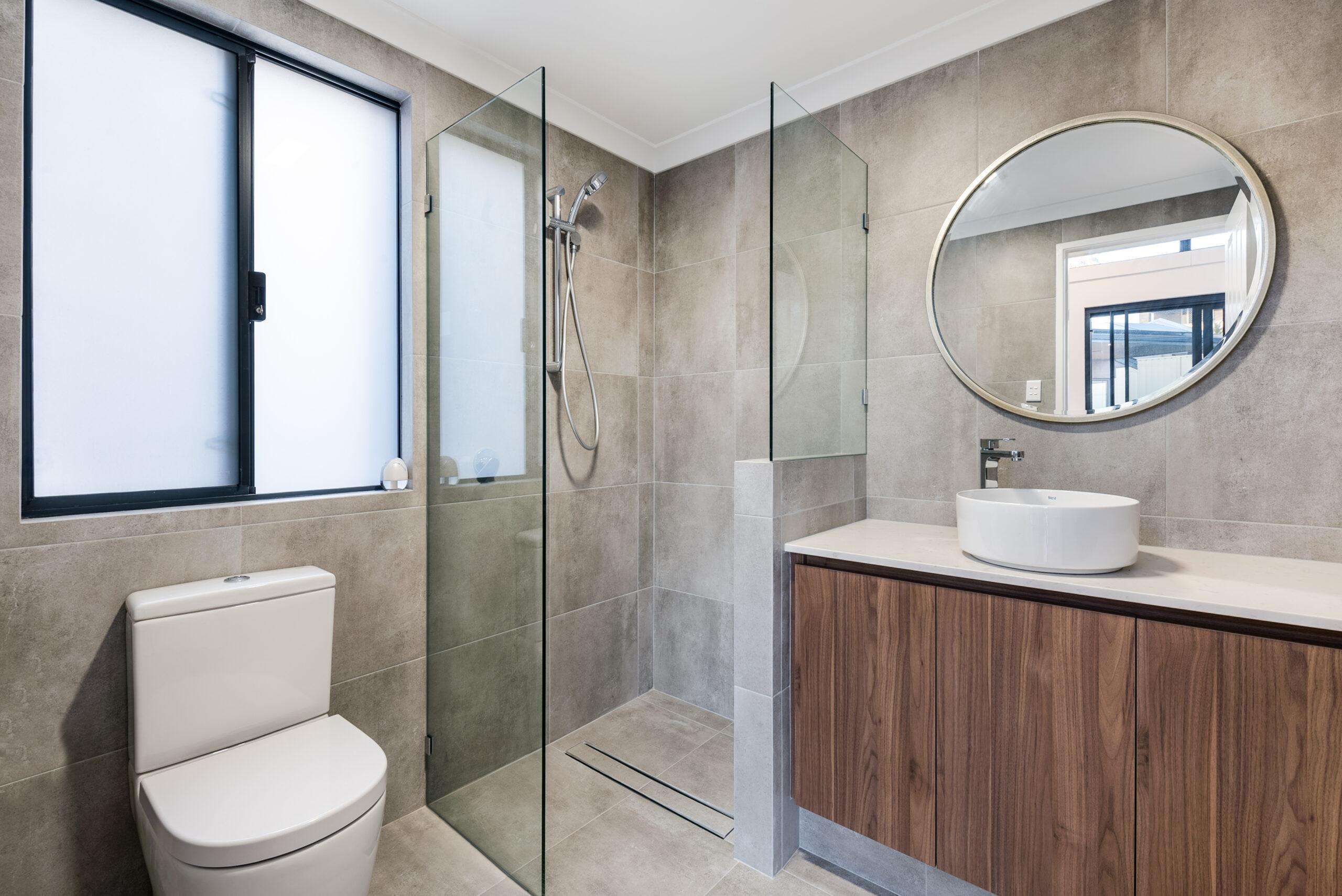 A modern bathroom in this Quinns Rocks granny flat boasts a glass-enclosed shower, round mirror, and a white sink atop a wooden vanity. Beige tiles adorn the walls and floor while a window floods the space with natural light.