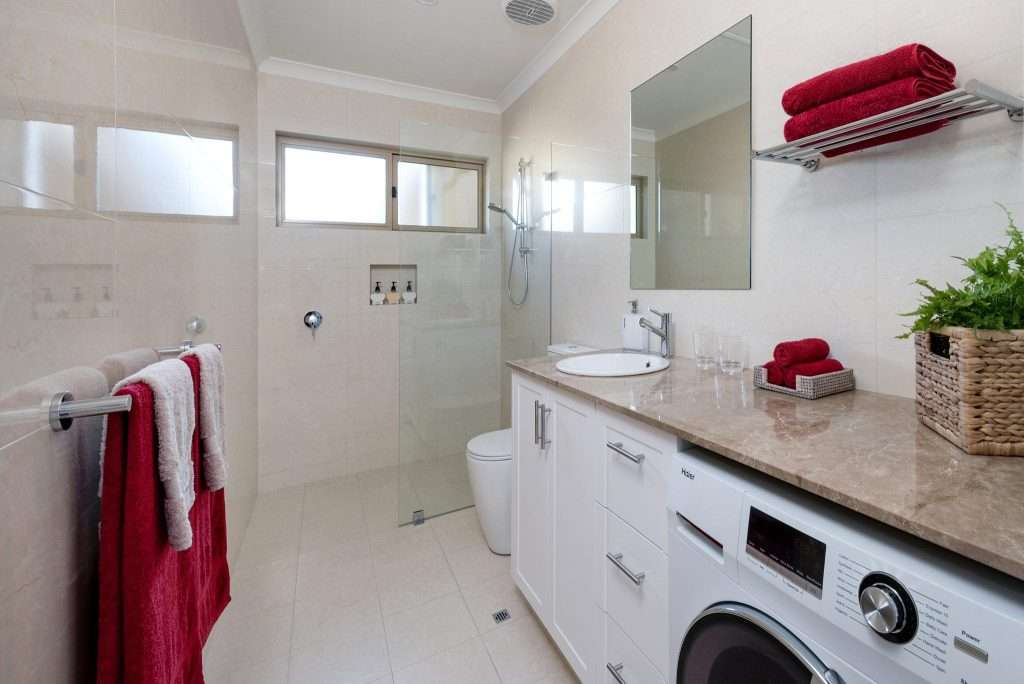 A sleek, modern bathroom in the Granny Flat features a shower, sink, and washing machine. Towels and a lively plant adorn the counter, complemented by pristine white cabinetry and elegant beige tiled walls and floor.