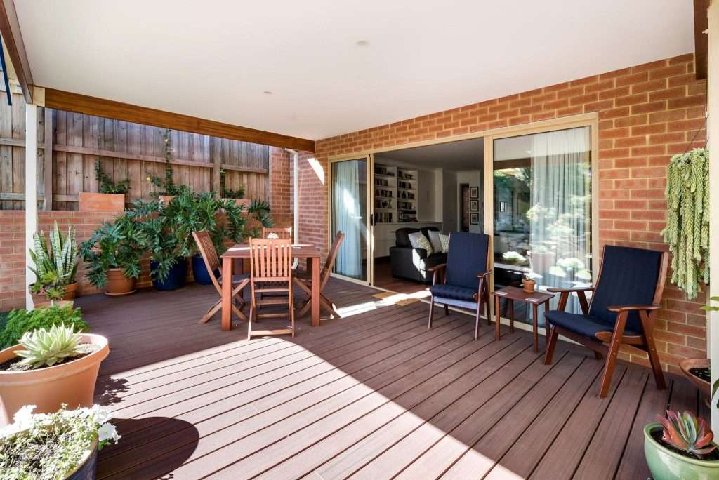 The Connolly Granny Flat boasts a charming covered patio with wooden flooring, complete with a dining table, chairs, vibrant potted plants, and two lounge chairs. Sliding glass doors invite you into the cozy living room adorned with bookshelves.