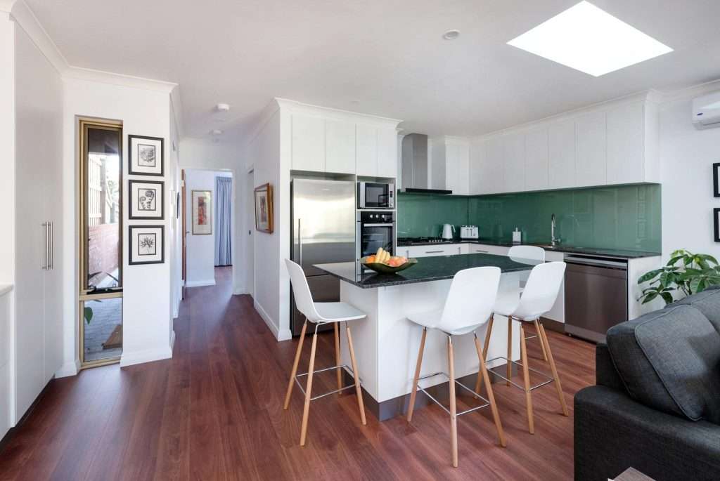 This modern kitchen in Connolly features white cabinets, a green backsplash, and a central island with bar stools. Hardwood floors seamlessly extend into the hallway, while a skylight above floods the space with natural light—perfect for any adjoining granny flat.