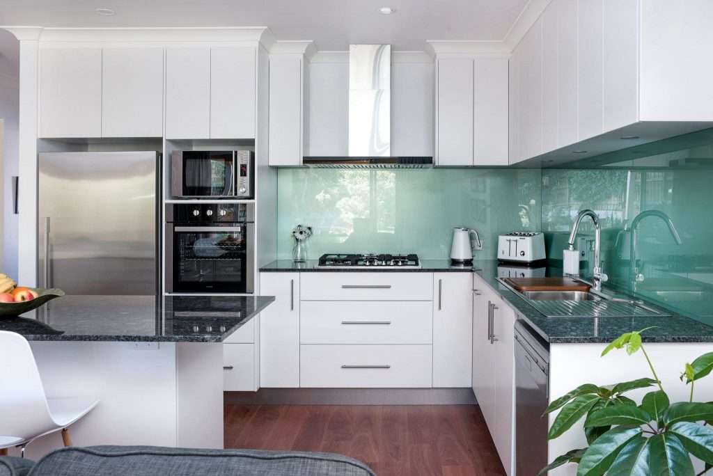 Modern kitchen in a Connolly home with white cabinets, stainless steel appliances, green glass backsplash, and wooden flooring. Features a stove, sink, fridge, and built-in oven. A cozy granny flat feel is enhanced by the houseplant in the corner.