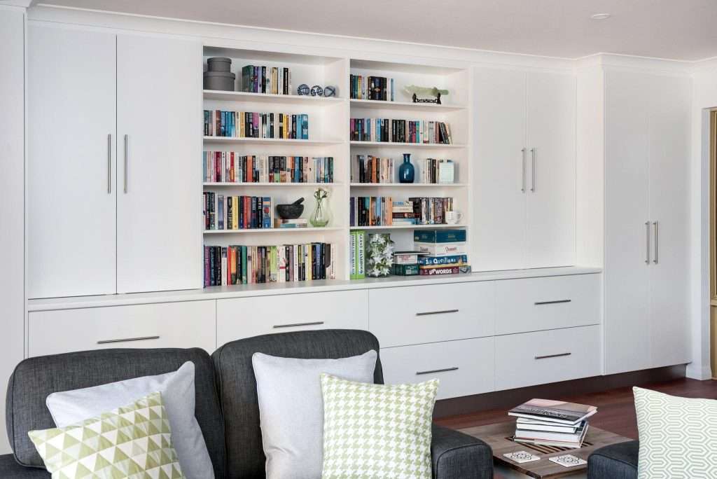 In a cozy granny flat, a white storage unit with shelves filled with books and decorative items is flanked by cabinets with silver handles. Two black Connolly chairs with patterned cushions sit in front, adding a touch of elegance.