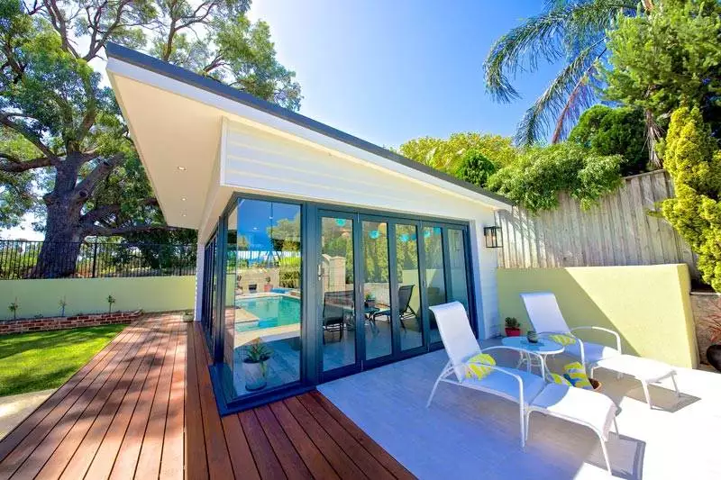 Modern poolside patio with glass doors, two white lounge chairs, a small table with two cups, and surrounding trees.