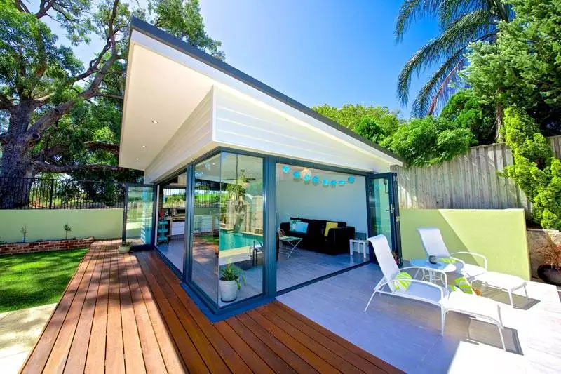 Modern pool house with glass walls, wooden deck, and white lounge chairs. Surrounded by trees and greenery under a clear blue sky.