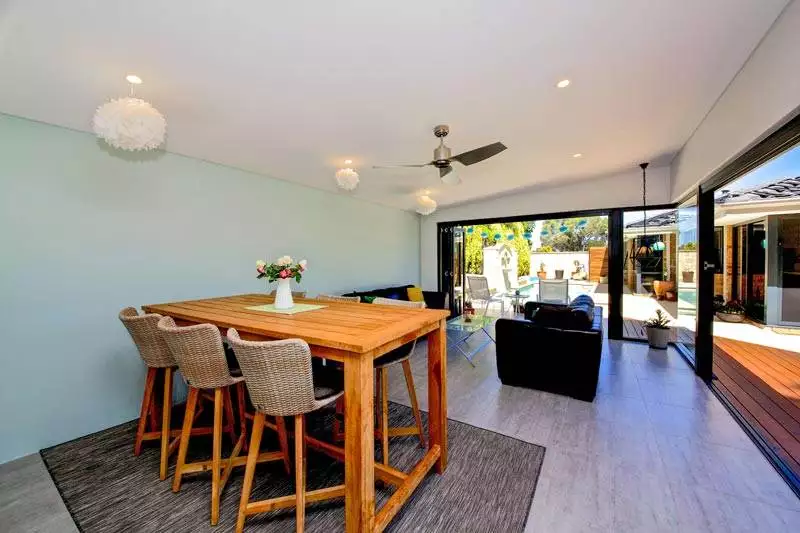 Modern open-plan dining and living area with a wooden table, wicker chairs, ceiling fan, and sliding glass doors leading to a patio and backyard. White hanging lights decorate the ceiling.